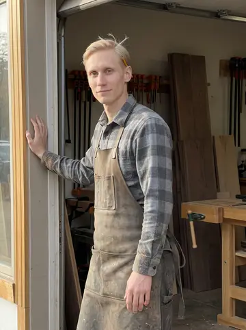 Per, AI character: the open doorway of a small Minneapolis cabinet shop, stacked walnut boards and clamps visible behind him