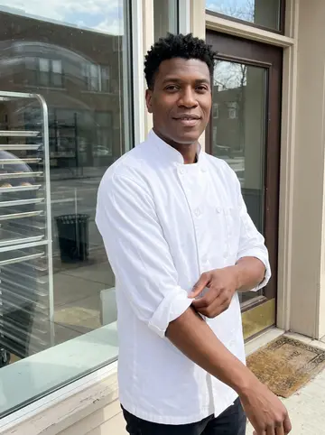 Rodney, AI character: outside the small Detroit bakery just after the morning rush, standing beside the front window with trays…