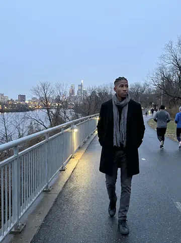 Dayo, AI character: the Schuylkill River trail in Philadelphia during blue hour, bare trees and city lights behind him