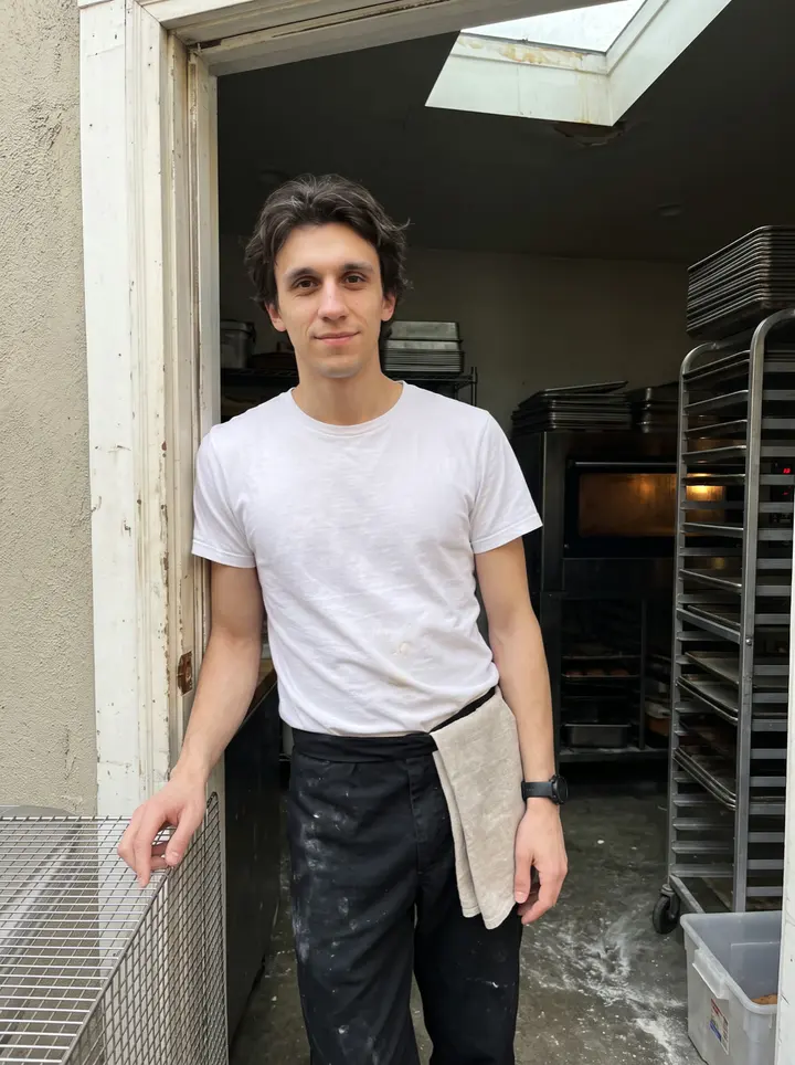 Fabien, AI character: the open back doorway of the Plateau bakery after the morning bake, metal racks and a leaking skylight…