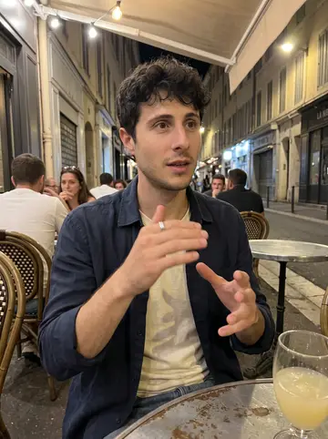 Yassine, AI character: a small terrace café in Marseille after sunset, pastis glasses and metal tables around him