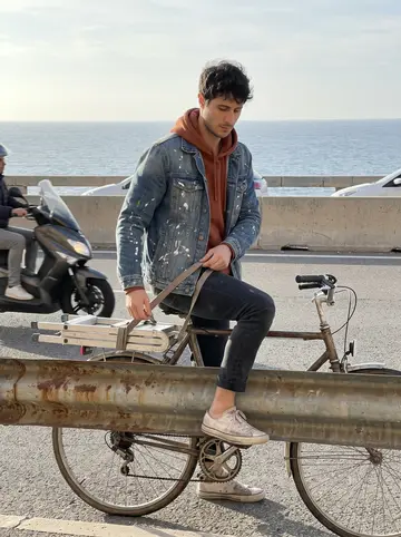 Yassine, AI character: the Corniche in Marseille with the sea behind him and a folded ladder strapped to the back of his old bicycle