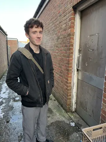 Jordan, AI character: outside the hotel kitchen service entrance in Dorchester after an early shift, leaning against a brick…