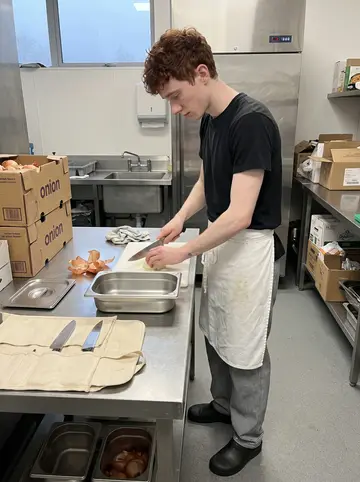 Jordan, AI character: quiet hotel prep kitchen before breakfast service, stainless prep table, onion boxes and a canvas knife…