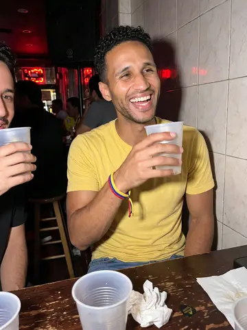 Yeferson, AI character: a small Colombian bar in Jackson Heights after a late gig, bandmates partly cropped at the table