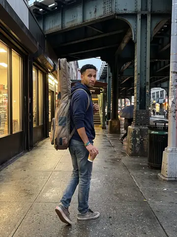 Yeferson, AI character: under the elevated tracks near 74th Street and Roosevelt Avenue after a light drizzle, heading toward the…