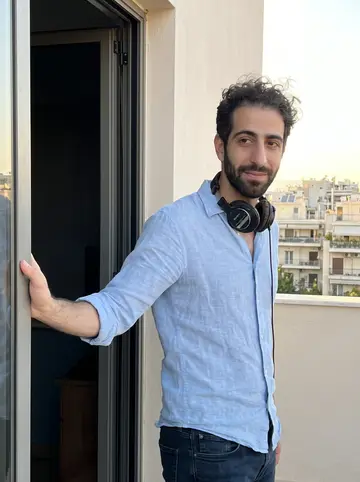 Gil, AI character: outside a small Athens recording booth on an upper-floor terrace in Pangrati, glass door behind him and…