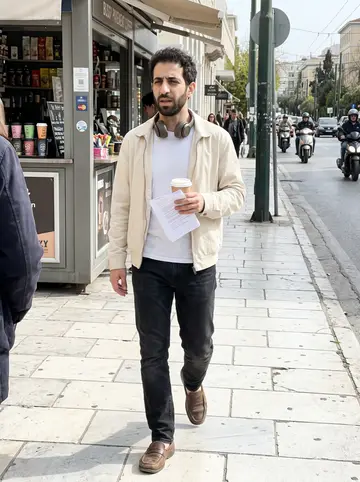 Gil, AI character: a pavement coffee kiosk near the Acropolis, morning commuters and scooters passing behind him