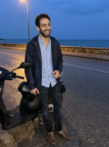 Gil, AI character: the coastal road near Vouliagmeni after a light drizzle, his dark moped parked beside a low wall with the…