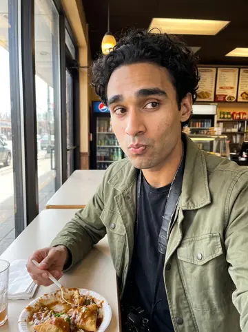 Faisal, AI character: a Devon Avenue snack counter in Chicago, paper plate of samosa chaat and a pint glass pushed near the edge…
