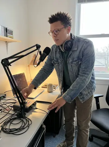 Reuben, AI character: inside the podcast office edit bay, standing beside a desk covered with a laptop, audio interface, cables,…