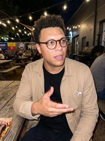 Reuben, AI character: a crowded Austin barbecue bar patio at night, neon beer signs and picnic tables behind him