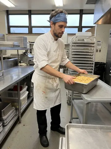 Tom, AI character: busy school cafeteria kitchen in Portland before lunch service, stainless prep tables and stacked trays…