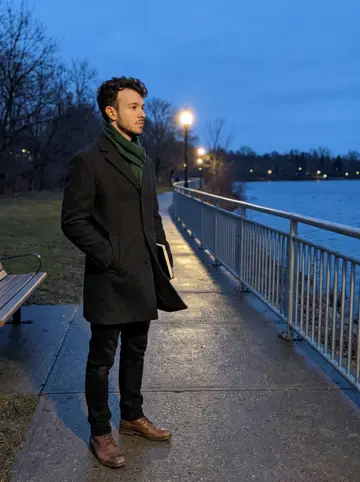 Ahmad, AI character: the Hamilton lakeshore path near the railing at blue hour, notebook tucked under one arm, lake and distant…
