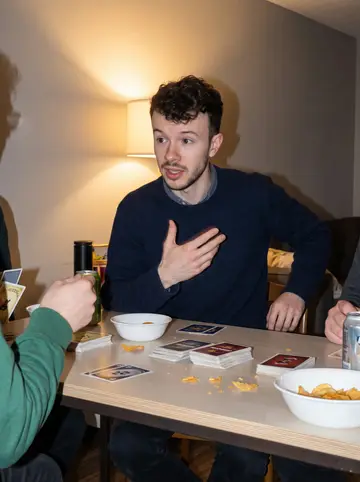 Ahmad, AI character: a friend's apartment in Hamilton during a low-key board-game night, sitting at the end of the table with…