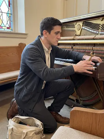 Alexei, AI character: a church sanctuary at 7am in Pittsburgh, kneeling beside an old upright piano with the front panel removed
