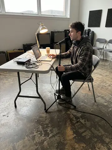 Tiernan, AI character: a borrowed rehearsal room with a folding table, microphone stand, laptop, coffee cups and guitar amps…