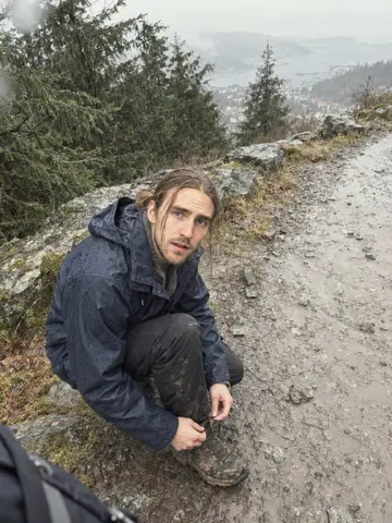 Erik, AI character: on the muddy Fløyen trail above Bergen on a rainy Saturday morning, wet spruce and city rooftops far below