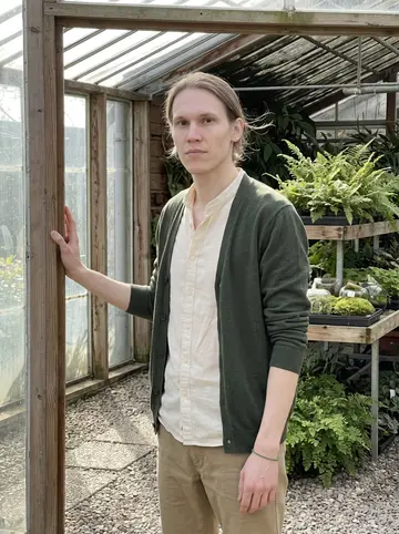 Tijmen, AI character: the open doorway of a glass greenhouse at his Rotterdam garden centre, trays of ferns and moss terrariums…
