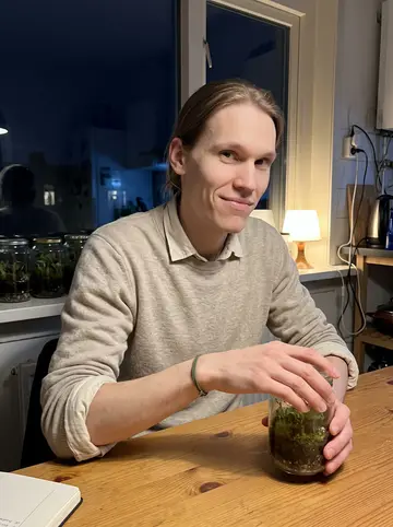 Tijmen, AI character: his Rotterdam apartment kitchen table late at night, old jam jars with moss terrariums lined along the…
