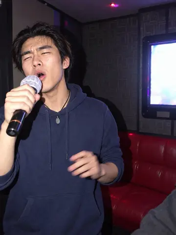 Shin, AI character: in a private noraebang room on Kingsway on a Thursday night, red vinyl bench and a lyric screen behind him