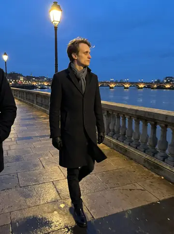 Jérôme, AI character: on the Garonne quays at dusk, stone railing and dark river behind him with Bordeaux lights starting to come on
