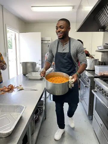 Babatunde, AI character: his mother's church hall kitchen during a Sunday fundraiser in Decatur, big pots and foil trays on a prep…