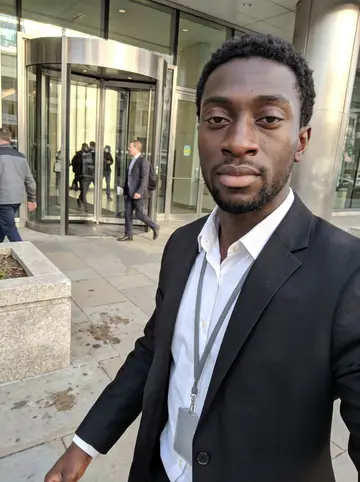 Ifeanyi, AI character: outside the glass tower downtown after his lobby shift, leaning against a stone planter near the revolving…