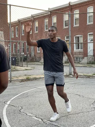 Khalil, AI character: cracked outdoor basketball court in West Baltimore, chain-link fence and row houses behind him