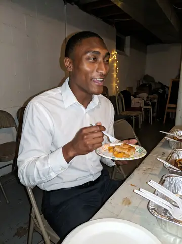 Khalil, AI character: church basement after Sunday service, folding tables, foil trays, and warm holiday string lights in the…