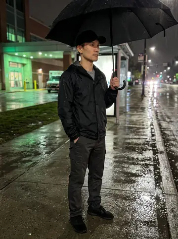 Haruki, AI character: bus stop outside the hospital during a light spring rain, wet pavement reflecting ambulance bay lights