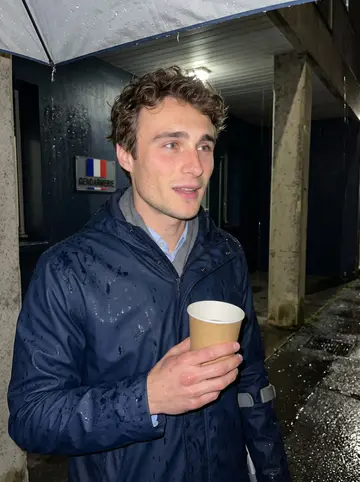 Marius, AI character: outside the local gendarmerie building during a rainy evening visit, under the covered entrance with wet…