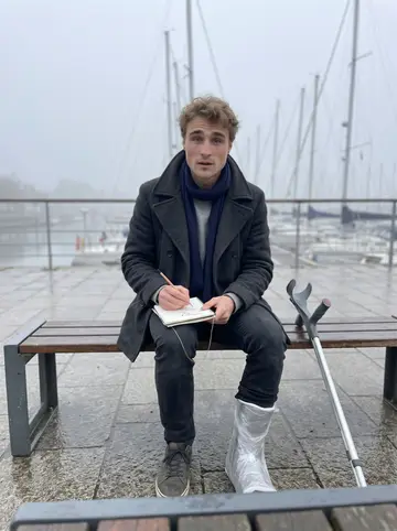 Marius, AI character: a harbor bench in Quimper in pale morning fog, boats and wet stone behind him, his worn pocket notebook…