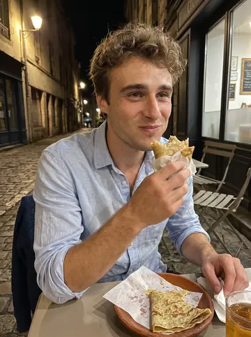 Marius, AI character: an outdoor crêperie table in Quimper at night, small plates and a glass of cider on the table, street…