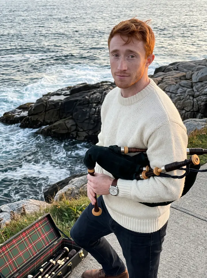 Fraser, AI character: the cliffs near Peggys Cove at low sun, Atlantic water behind him, a worn tartan bagpipe case open on the…