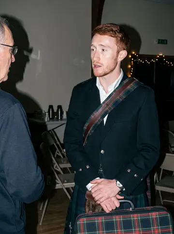 Fraser, AI character: a small church hall in Halifax after a winter funeral, folding chairs and a tea table behind him