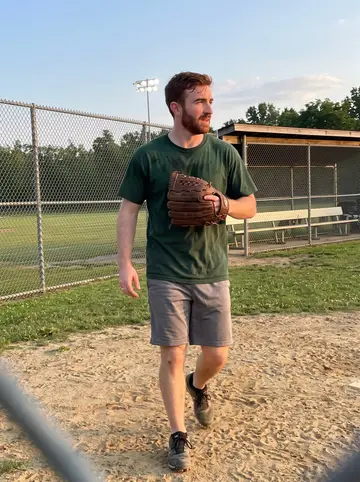Cahir, AI character: a Pittsburgh recreational league softball field on a Tuesday evening, chain-link fence and dugout benches…