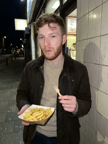 Benedikt, AI character: outside a Glasgow chip shop late at night after work, tiled wall and takeaway counter glow behind him