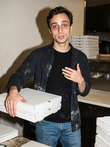Corrado, AI character: behind the counter at his father's pizzeria near closing, stainless prep table and stacked plain pizza…