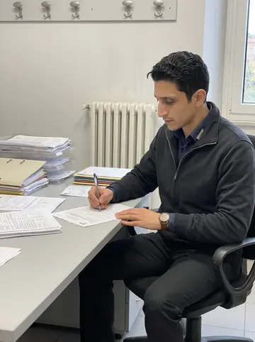 Stelvio, AI character: a plain office corner at the carabinieri station in Turin, stacks of forms on a grey desk, coat hooks and…