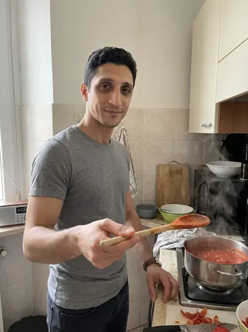 Stelvio, AI character: his small Turin kitchen on a Sunday morning, tomato sauce pot on the stove, radio and cutting board on the…