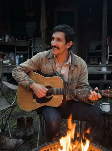 Esteban, AI character: the bunkhouse fire pit at dusk, a worn acoustic guitar on his knee, the dark outline of the bunkhouse…