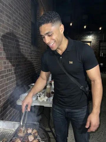 Isaac, AI character: the courtyard of his Bronx apartment building during a humid summer night cookout, folding table and foil…