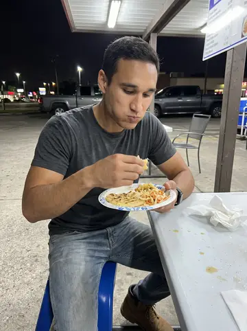 Ernesto, AI character: a pupusa stand parking lot in Houston after midnight, plastic tables under a strip-mall awning