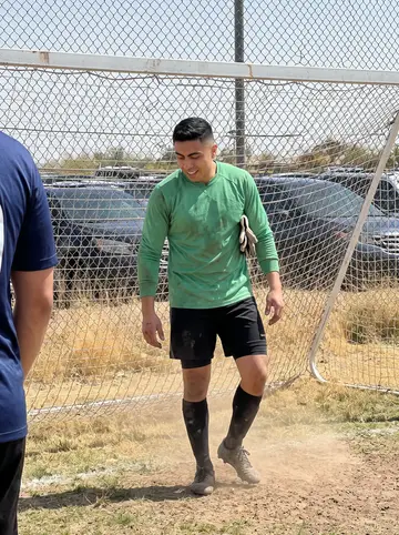 Eduardo, AI character: a dusty Saturday pickup soccer field on the edge of Phoenix, chain-link fence and parked cars beyond the goal