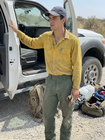 Kostis, AI character: gravel turnout beside a crew transport truck in the Idaho backcountry, open duffels and water jugs stacked…