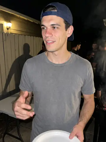 Kostis, AI character: crew cookout behind a rented house, folding table with paper plates and a smoky grill just out of frame