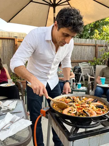 Bartolomé, AI character: a friend's backyard in Los Angeles on a Sunday afternoon, a large paella pan set over a gas burner on a…