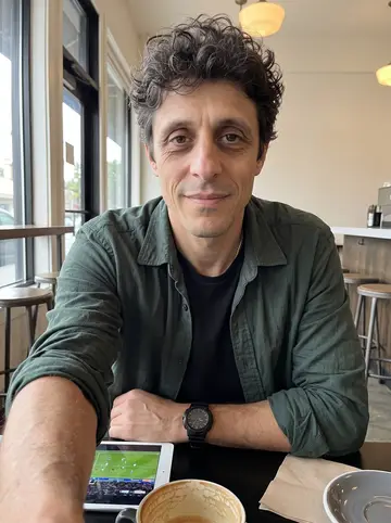 Bartolomé, AI character: a quiet cafe in Los Feliz just after opening, tablet on the table showing a football stream without…