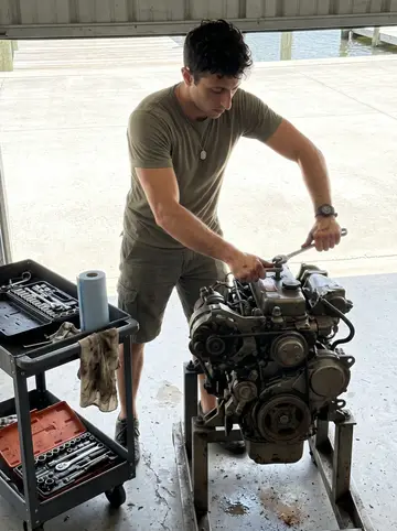 Fotis, AI character: open-sided marina repair bay in Tarpon Springs, tools on a rolling cart and an old Yanmar sailboat engine…